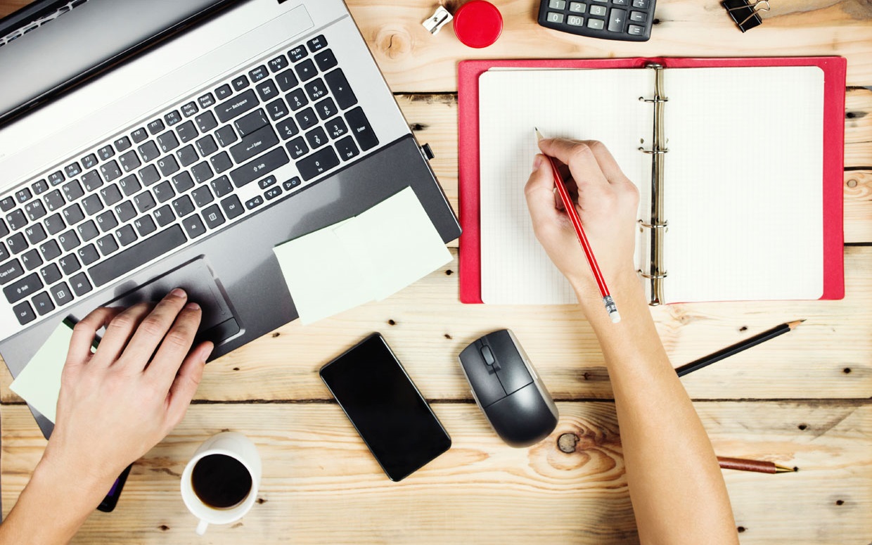 A image of a copywriter taking notes while browsing through his or her laptop, with a sharpener, calculator, and clip at the top of the image. The words "We Can Write Interesting and Informative Articles for Your Website!" is in the centre of this photo.